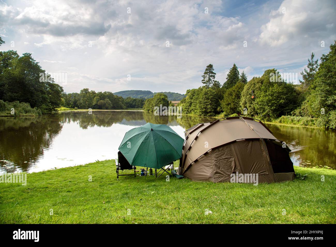 A tent and large umbrella stand on the bank of a lake surrounded by beautiful trees. A peaceful location to relax and enjoy the great outdoors. Stock Photo