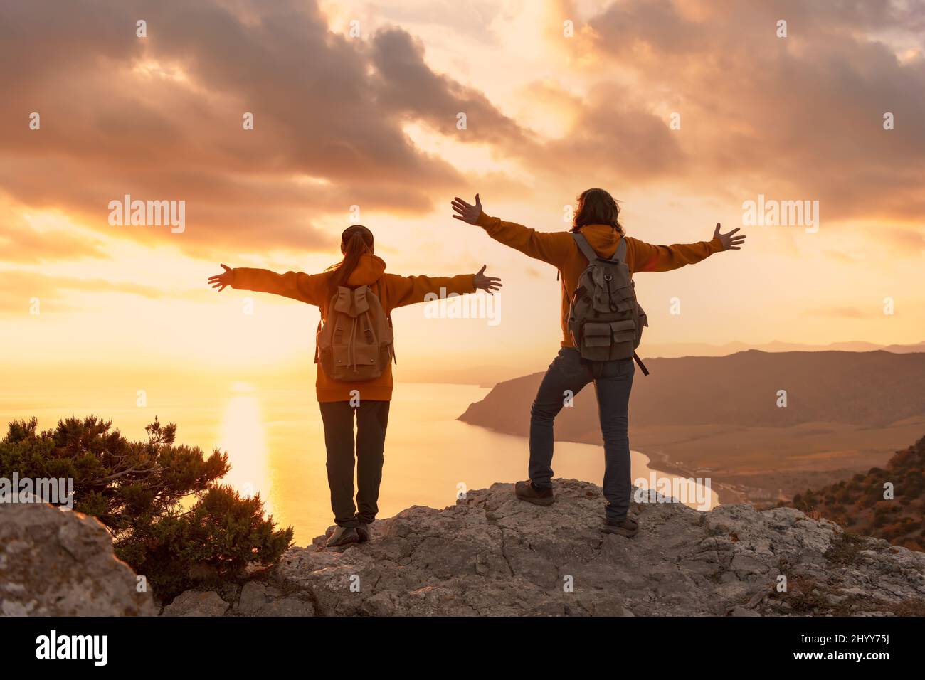 Couple of two young hikers stands with raised arms at mountain top and looks at sea and sunset Stock Photo