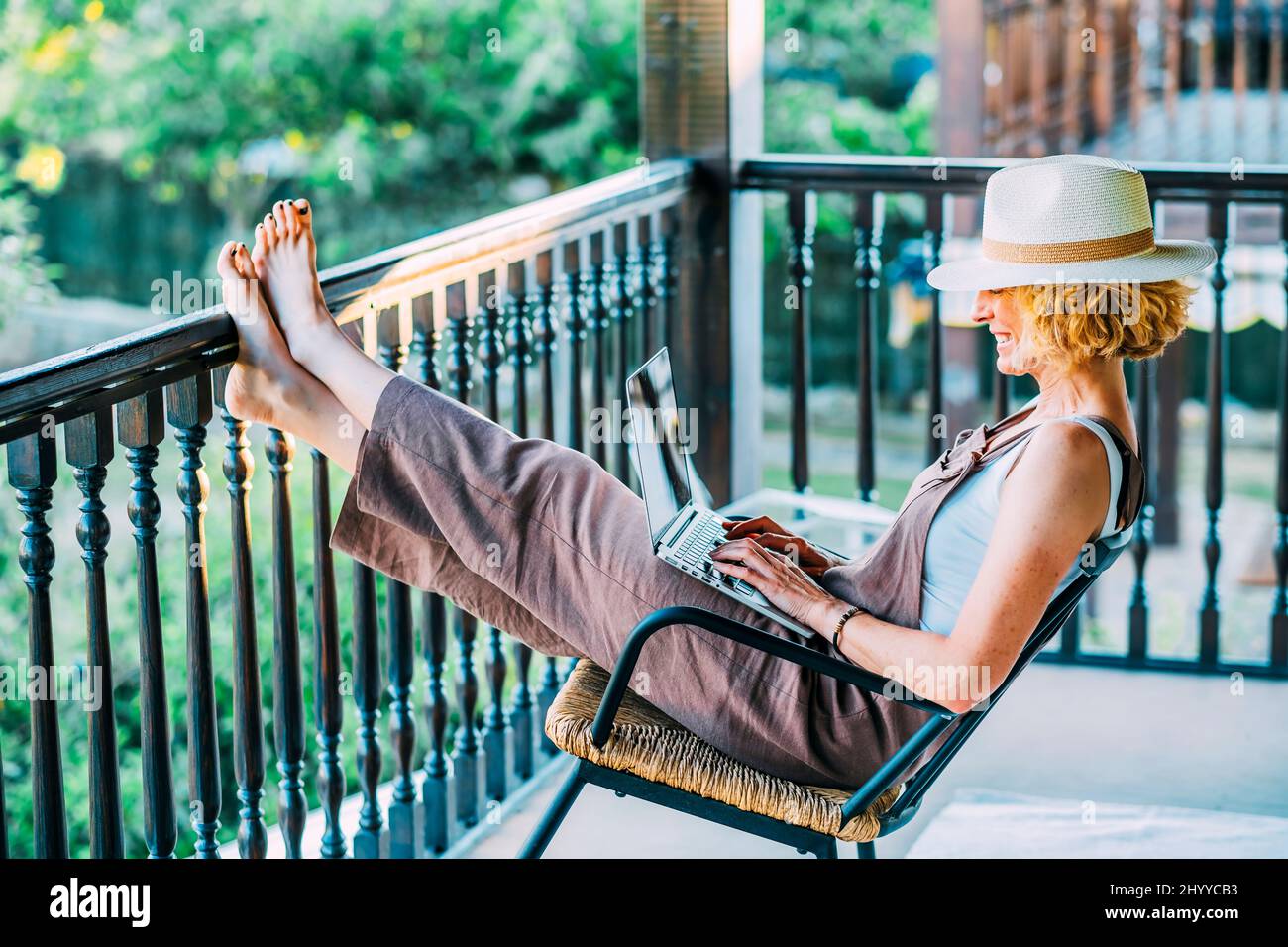 Portrait of a mature caucasian blonde young woman in her 50s with a laptop wearing a hat and sitting in a relaxed pose in a rocking chair on the woode Stock Photo