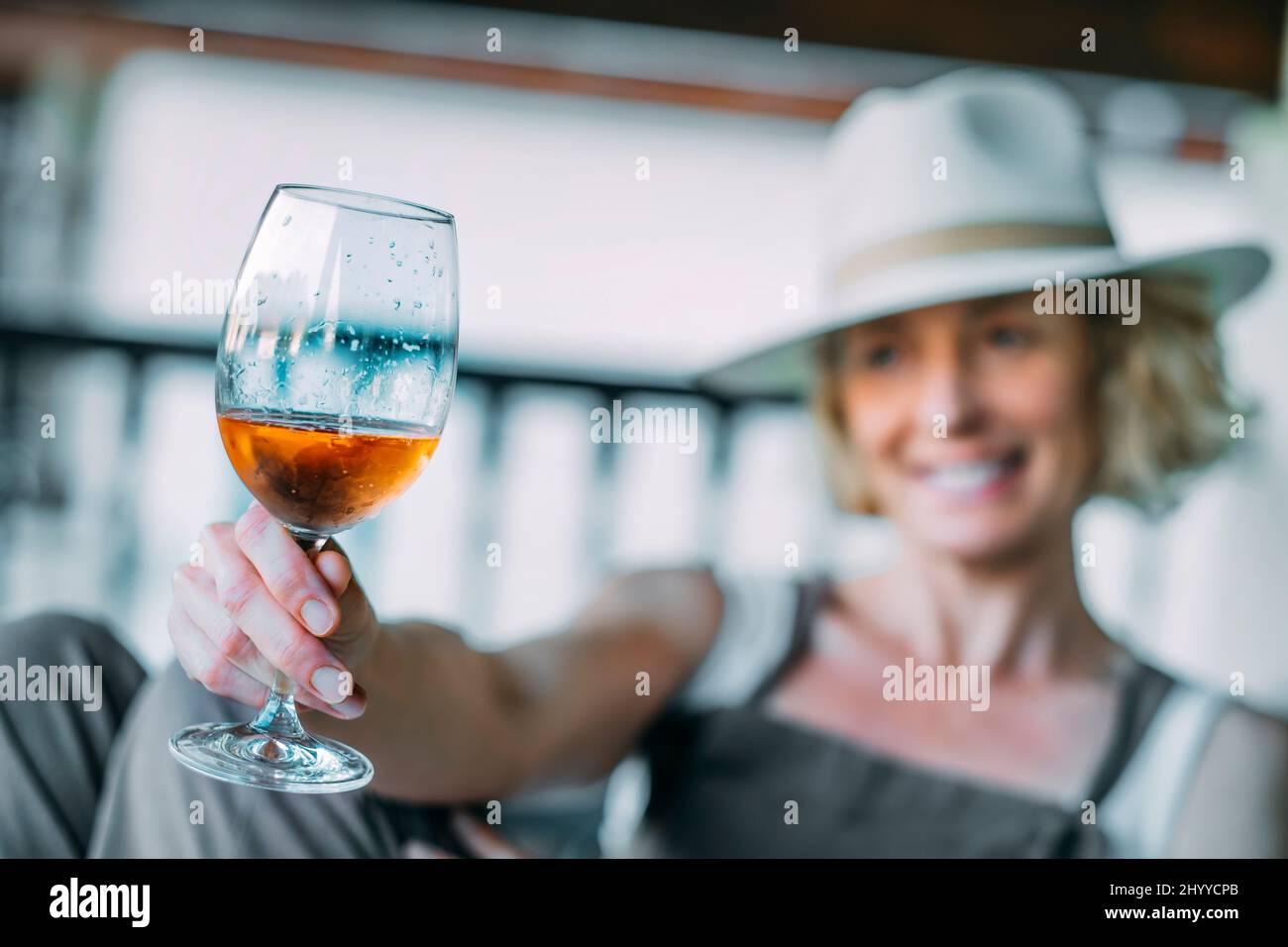 Portrait of a mature caucasian blonde young woman in her 50s  wearing a hat and  enjoying a glass of wine on the  porch of a country house. Lifestyle Stock Photo