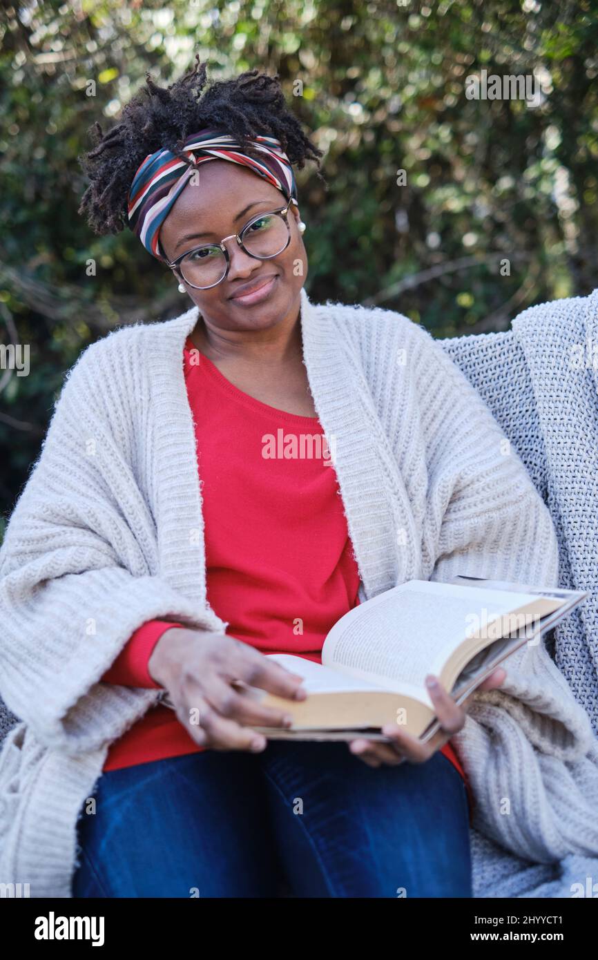 Portrait of a young black woman with afro hairstyle and glasses reading a book sitting on a garden old bench. Lifestyle concept. Stock Photo