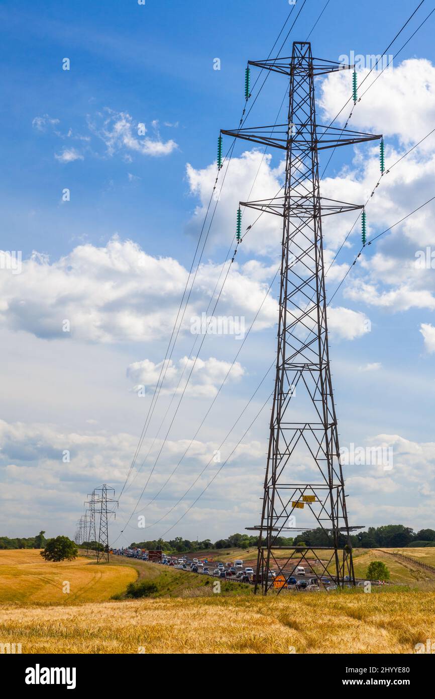 Electrical pylon power lines and traffic cars queuing on a motorway or freeway with blue sky clouds and sunshine Stock Photo