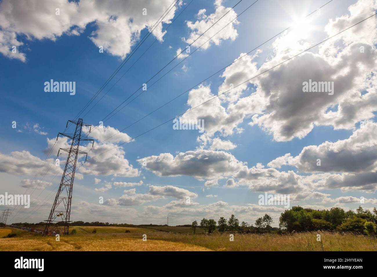 Electrical pylon power lines and traffic cars queuing on a motorway or freeway with blue sky clouds and sunshine Stock Photo