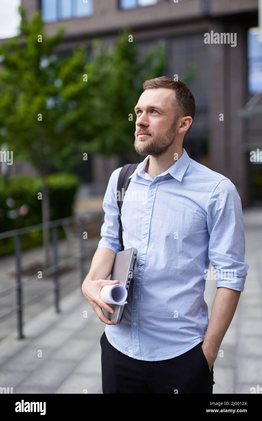 Young stylish serious bearded business man standing on a street with laptop and blueprints. Portrait of successful and confident businessman, designer or architecture expert. High quality image Stock Photo