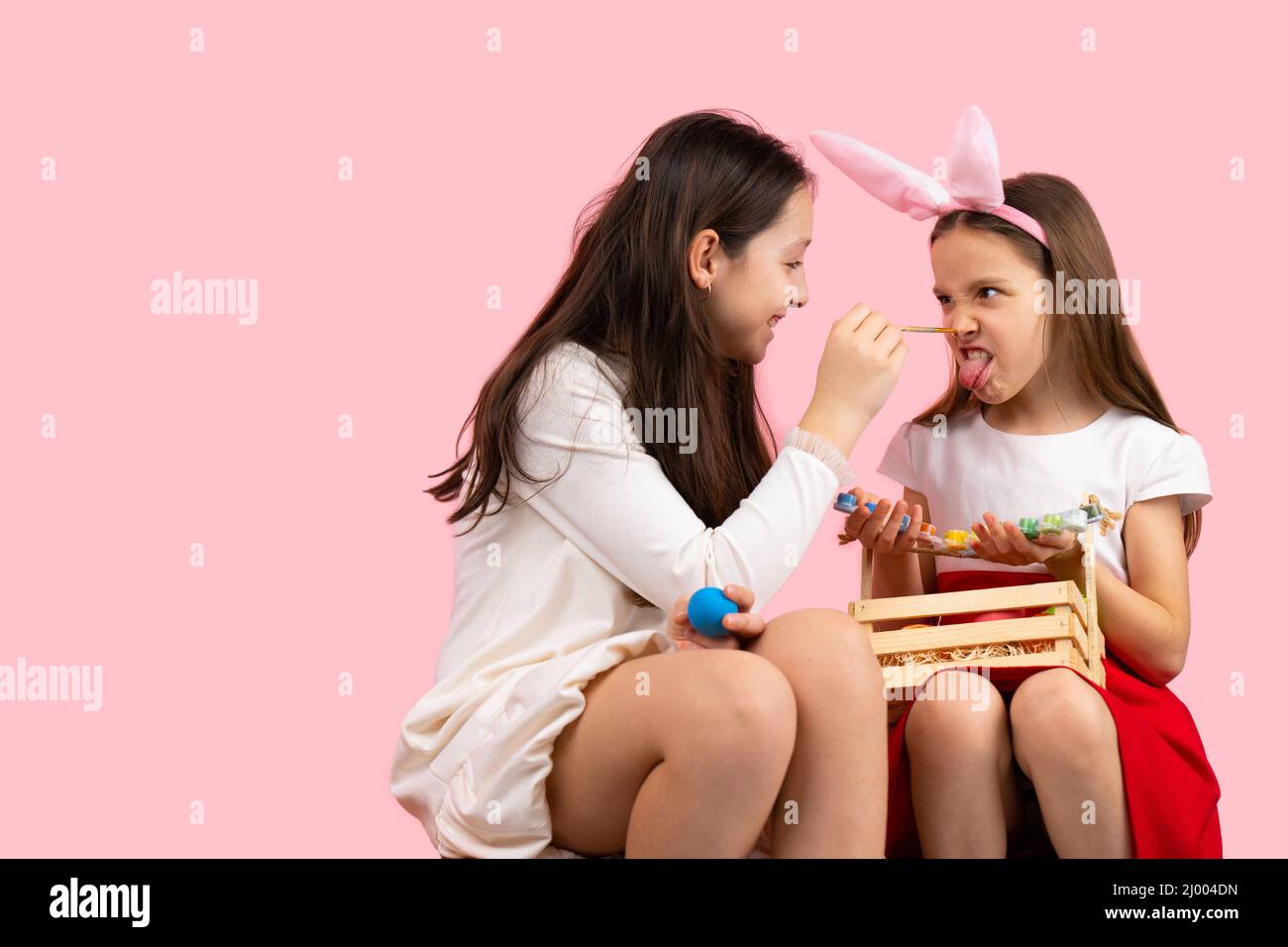 Picture of two sisters preparing for the Easter party wearing bunny ears and painting the nose, making fun together. Stock Photo