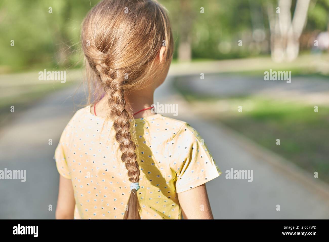 A little blonde girl in a yellow casual dress with a pigtail on her head, standing with her back to the camera. High quality photo Stock Photo