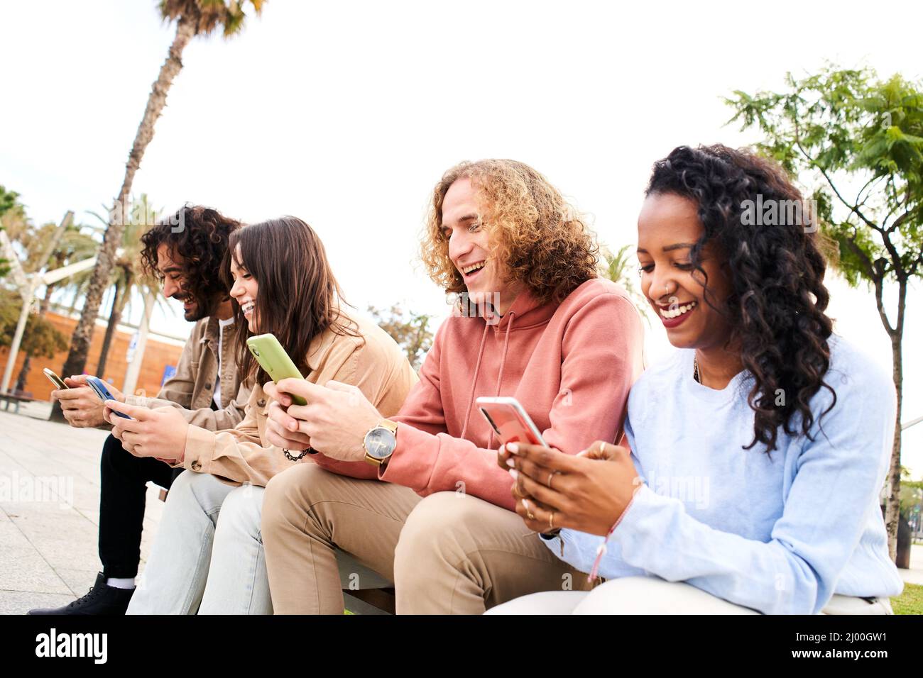 Friends having fun sharing in social media. Smiling mixed race group of friends sitting together on bench using mobile phone in the park Stock Photo