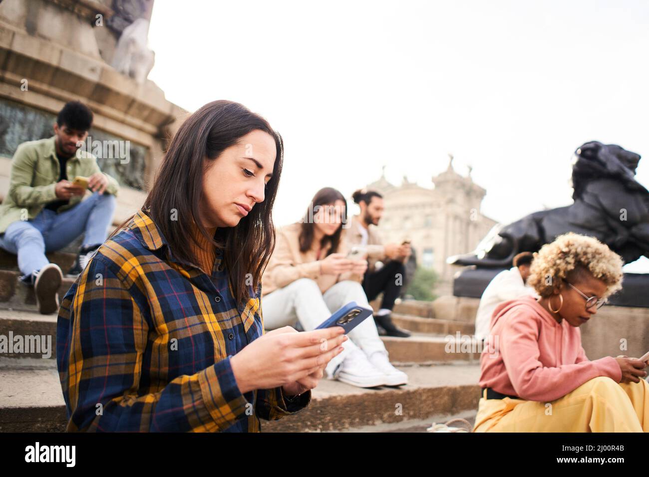 Group of individualistic people using phone outdoors with serious face. Friends focused on their mobiles sitting on a staircase in a city. Technology Stock Photo