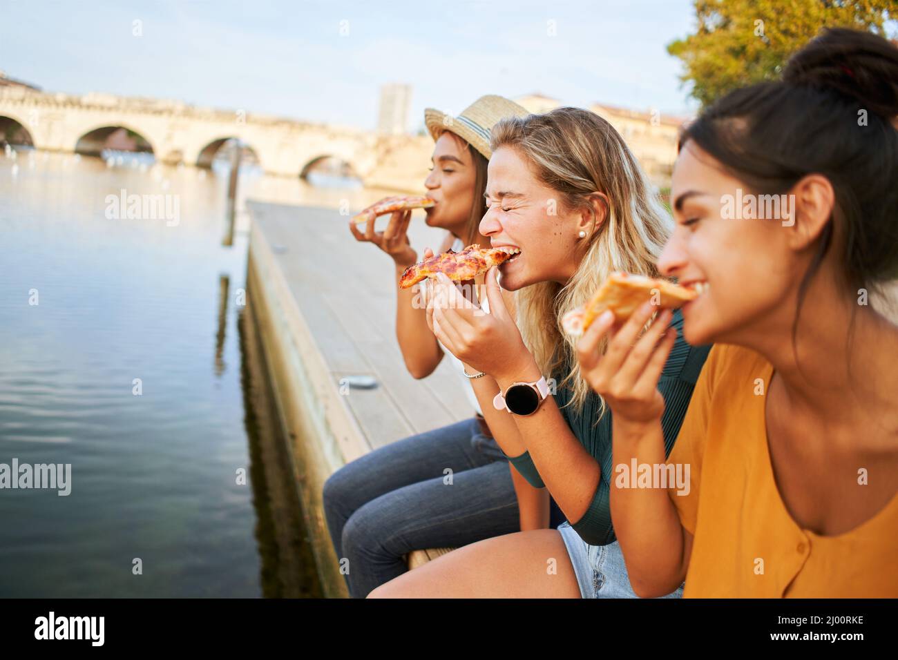 Three happy women sitting on the lakeside of the city eating pizza in a street stall. The happy girls enjoy the weekend together outdoors. Stock Photo