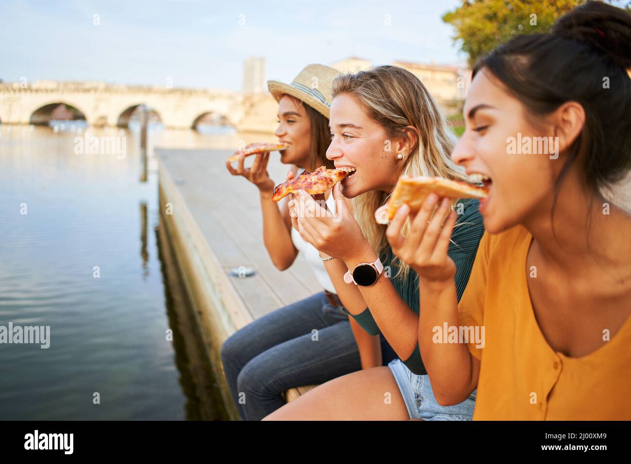 Three beautiful girls eat pizza on the lakeside. Group of women enjoy free time together sightseeing in a roman city in europe. Stock Photo