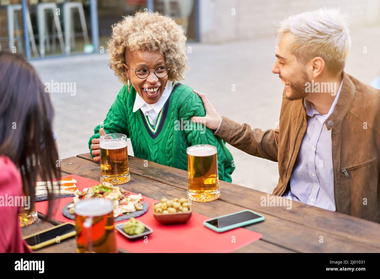 Multiracial couple having fun. Happy people celebrating drinking beer together with friends at terrace bar. Man and woman laughing outdoors. Stock Photo
