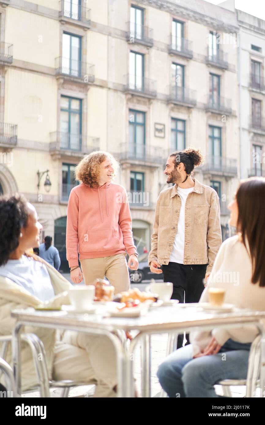 Vertical photo of Two female friends having breakfast on the terrace of the bar while 2 guys come over to flirt. Stock Photo