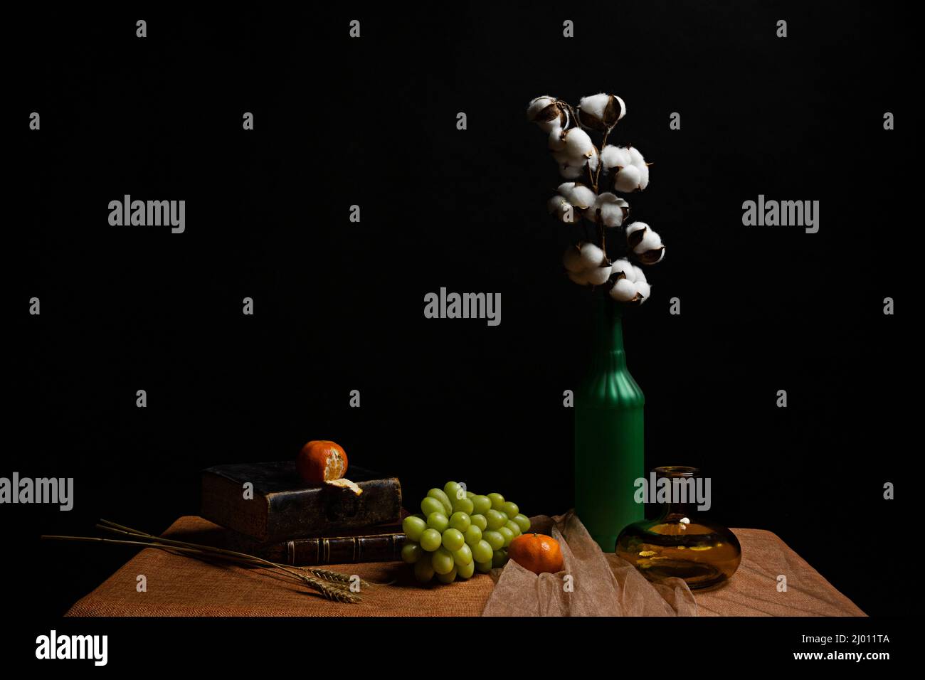 Still life with a cotton branch in a glass bottle with white grapes, tangerines, wheat branches, tulle fabric, glass jars and old books Stock Photo