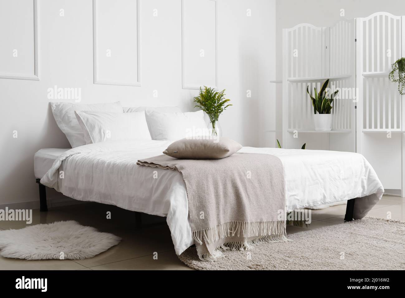 Comfortable bed and folding screen with houseplants near white wall