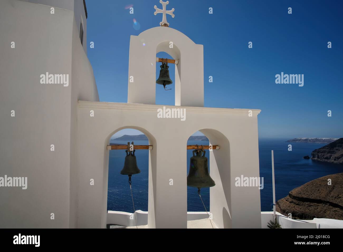 Three church bells and a religious cross on the top next to a church and a beautiful view of the aegean sea in Santorini Stock Photo