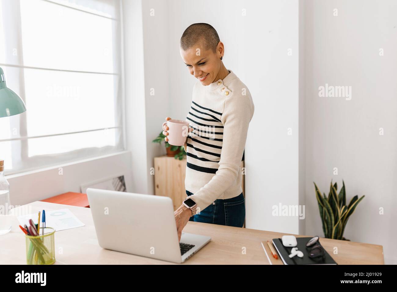 Mid adult businesswoman drinking coffee while using laptop at home office Stock Photo