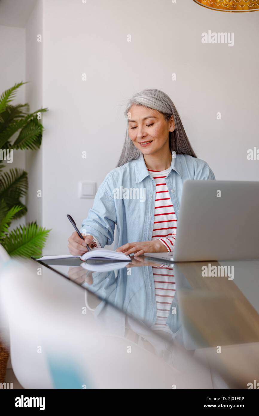 Joyful woman writing in notebook and using laptop at home Stock Photo