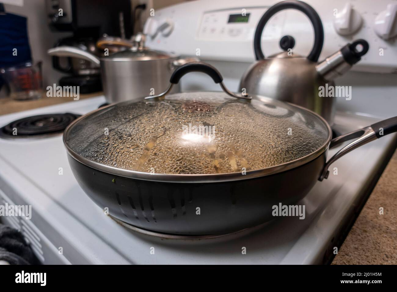Angled, selective focus of a large, covered skillet cooking dinner at a simmer on a white stove