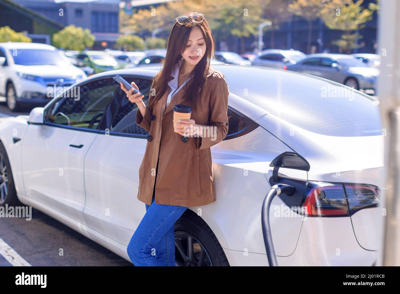 Smiling young  woman standing on city parking near electric car, charging automobile battery from small city station, drinking coffee and using smartp Stock Photo