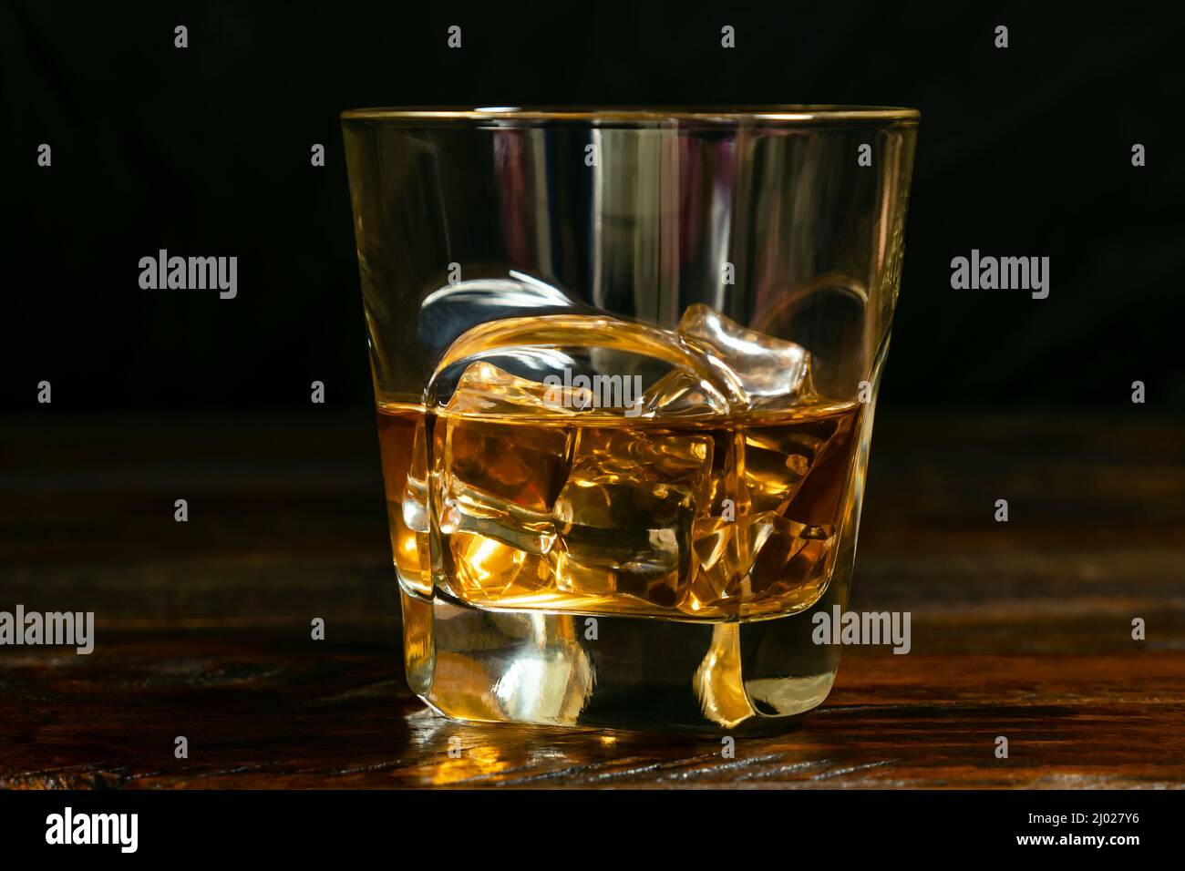 A close-up of a half-poured glass of whiskey with ice cubes on the dark oak boards Stock Photo