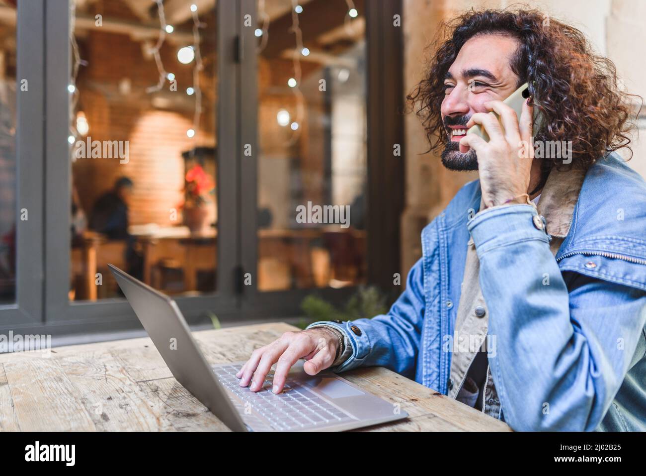 Gleeful Hispanic freelancer in casual clothes with curly hair smiling. and answering phone call while sitting at cafeteria table and using netbook on city street Stock Photo