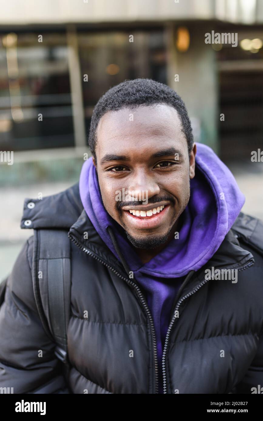 High angle of merry bearded black man in casual outerwear smiling and looking at camera on blurred background of city street Stock Photo