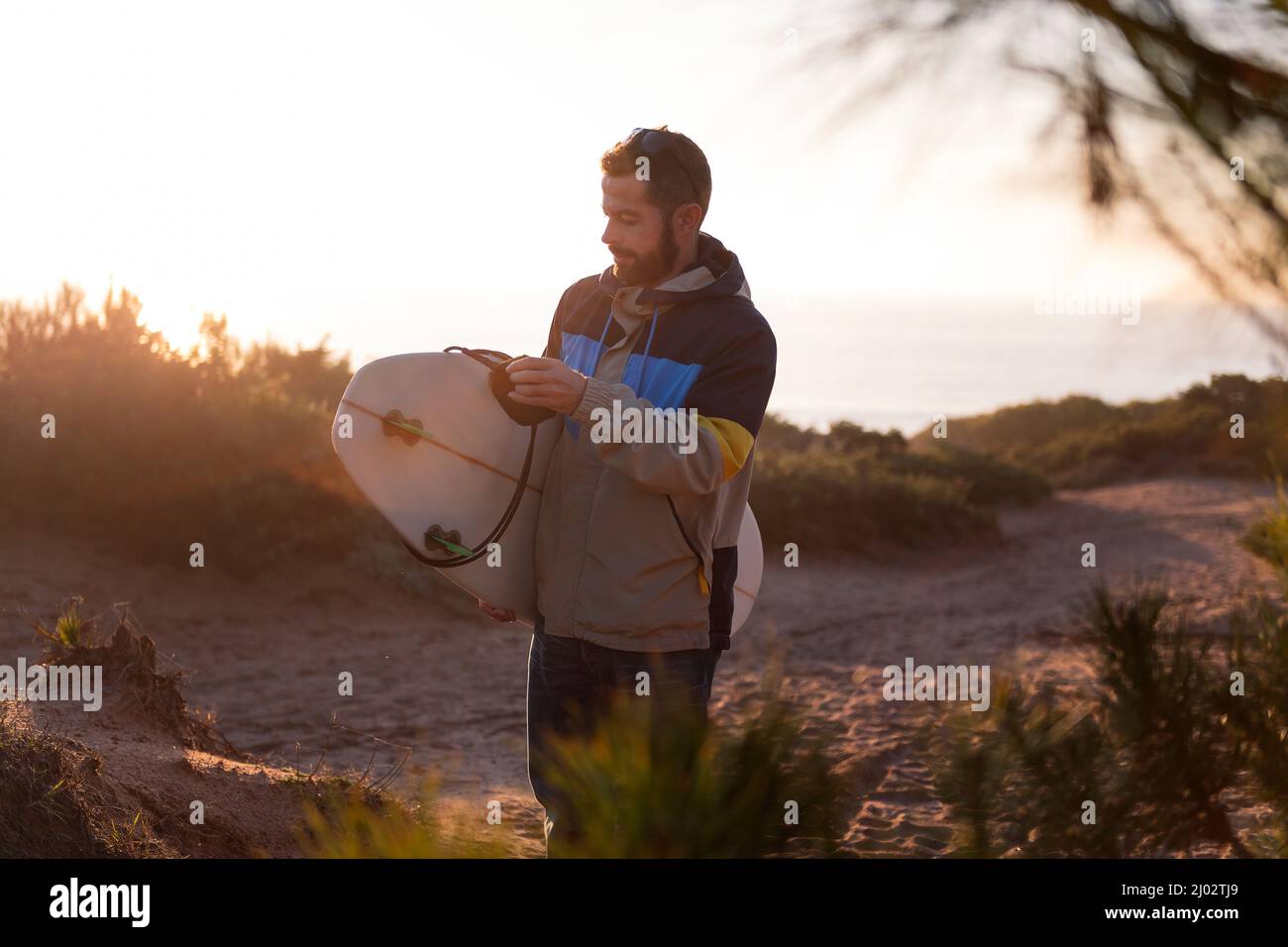 bearded young man in jacket walking at sunset by the dunes of the beach with his surfboard under his arm, leisure and hobbies concept, copy space for Stock Photo