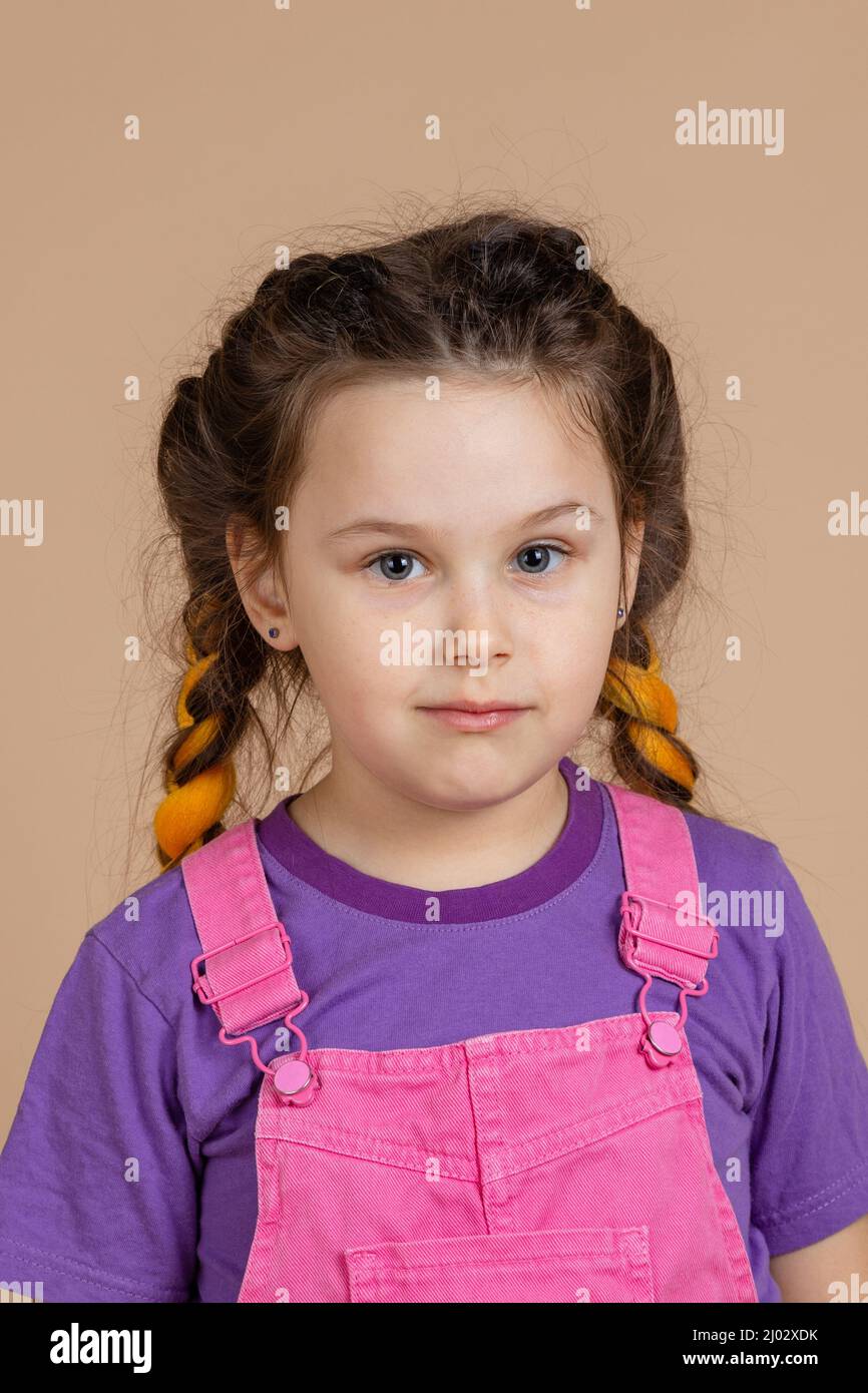 Portrait of pretty little girl with slight smile having yellow kanekalon braids, with eyes looking at camera wearing pink jumpsuit and purple t-shirt Stock Photo