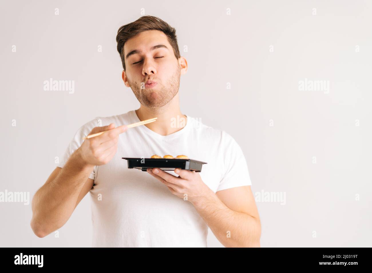 Portrait of handsome young man with enjoying eating fresh tasty sushi rolls with chopsticks on white isolated background. Stock Photo
