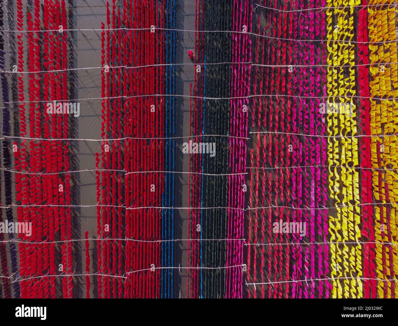 Narayanganj, Dhaka, Bangladesh. 16th Mar, 2022. Workers hang thousands of different colorful fabrics on iron wires tied between a bamboo framework and constantly turn them so that they dry perfectly in flooded field in Narayanganj, Bangladesh. Iron wires are used between a bamboo framework to create giant washing lines for the final part of the dying process as the fabrics are dried in the sun. Bright strands of blue, pink, orange and green-dyed cloths hang above the grassy field in a dazzling network of interlocking colors. This is the final part of the dying process after which the cloth is Stock Photo