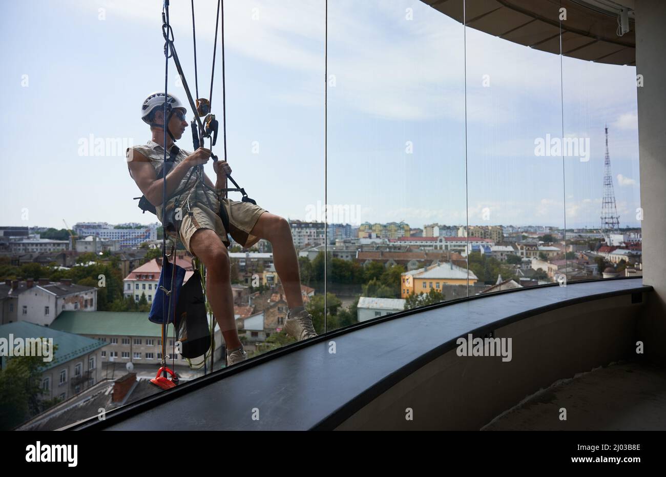 Industrial mountaineering worker hanging on rope and wiping window. View from inside building. Cleaner using safety lifting equipment while cleaning glass of high-rise building. Stock Photo