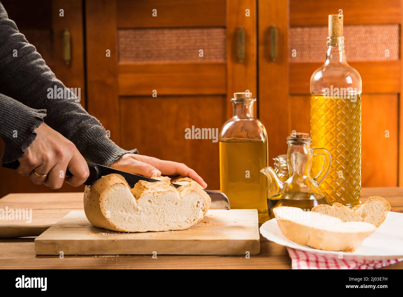Detail of hands with a knife cutting a slice of bread on a wooden board, next to extra virgin olive oil in several bottles and glass containers, on a Stock Photo