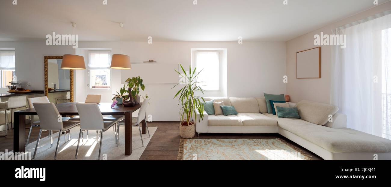 Front view of living room with a large sofa with cushions, table with leather chairs. Bright windows in a modern apartment. Nobody inside Stock Photo