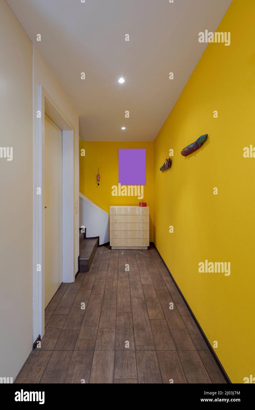 Front view corridor with yellow walls, there is a closed door and a chest of drawers at the bottom with brown stairs. Nobody inside Stock Photo