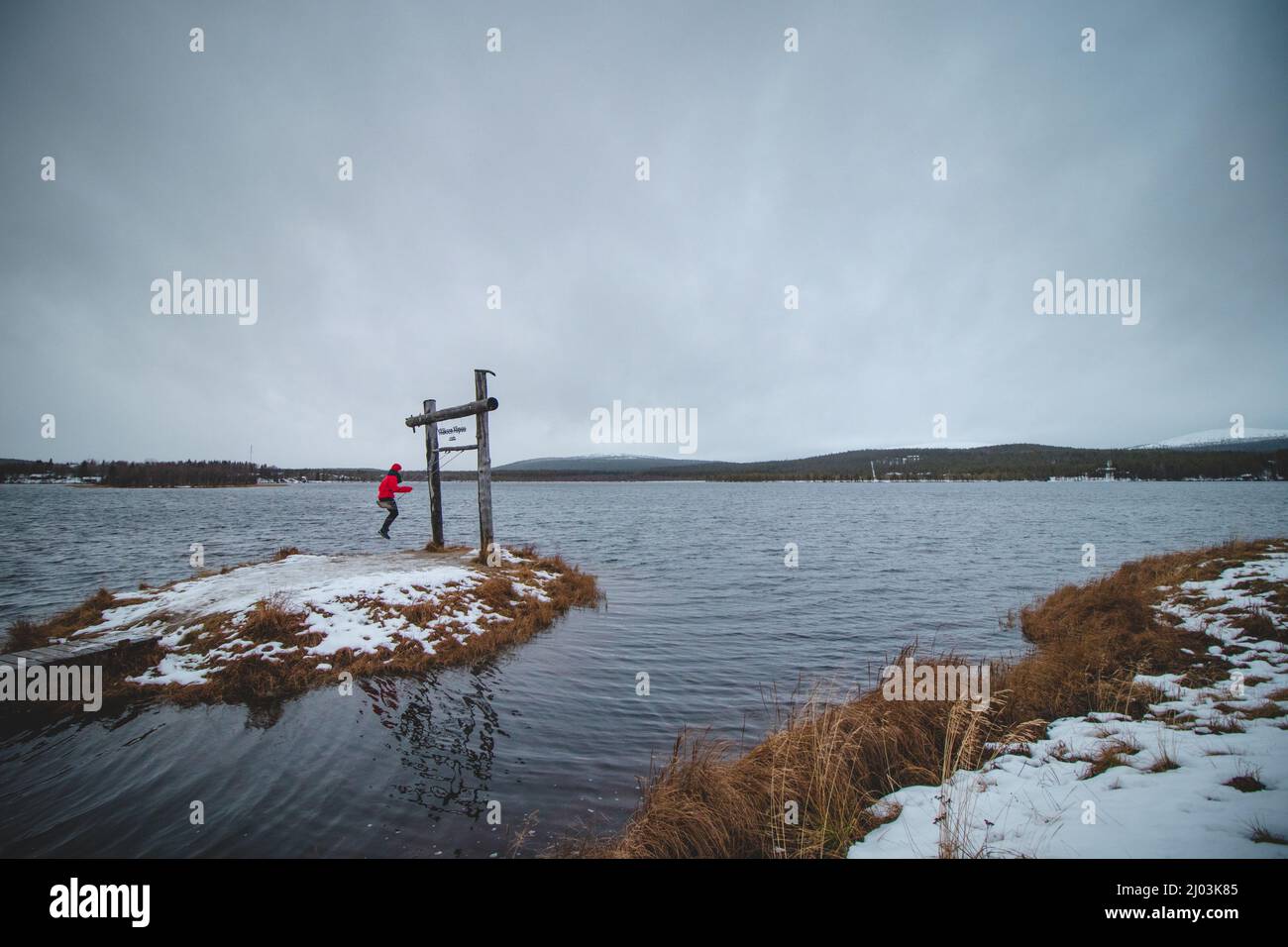 Woman in a red jacket enjoys a wooden swing in the town of Äkäslompolo in Lapland, Finland on a lake and the surrounding snowy mountains. The joy of a Stock Photo