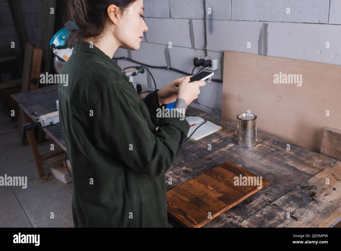 Side view of young craftswoman using smartphone near wooden plank in workshop Stock Photo