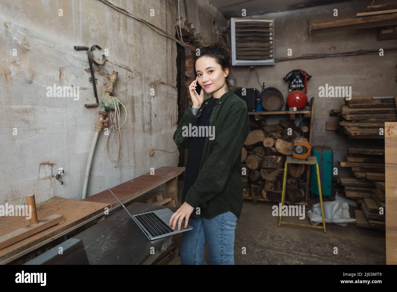 Smiling carpenter looking at camera while talking on smartphone and using laptop in workshop Stock Photo