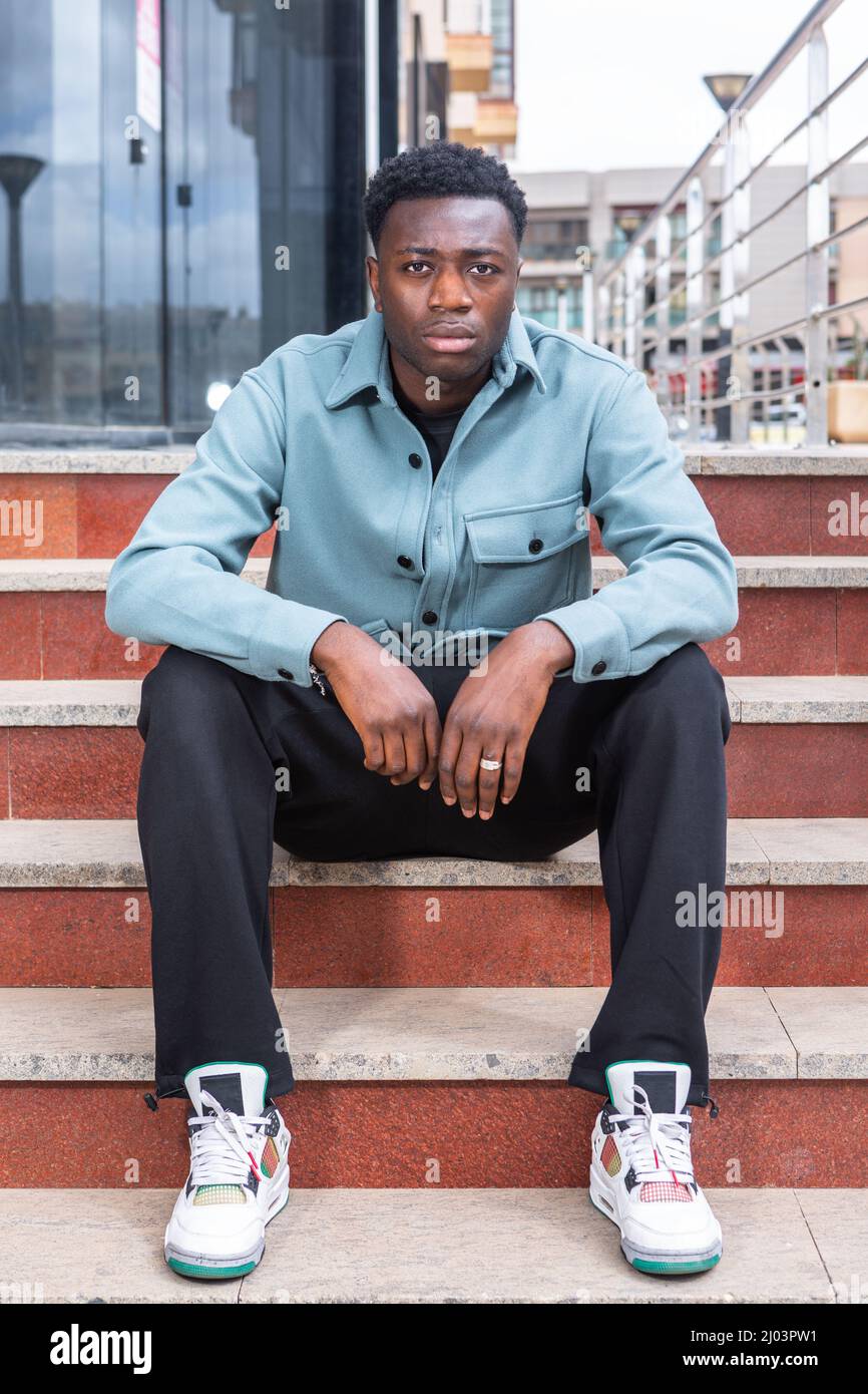 Full length of self assured young black man in trendy clothes resting on street stairs and looking at camera near modern building Stock Photo