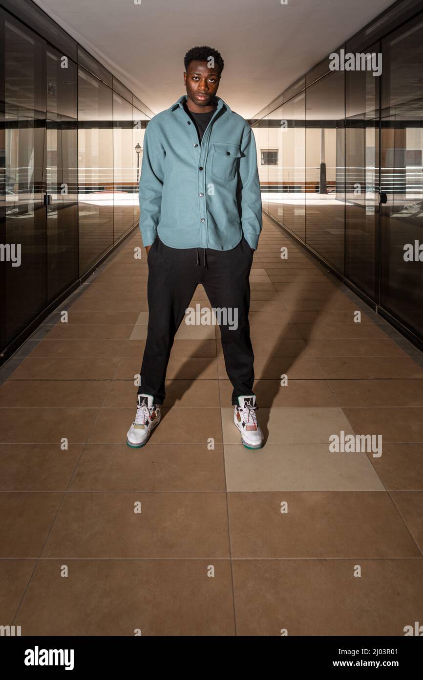 Full length of confident young black male in stylish clothes and sneakers looking at camera while standing in corridor of modern building with hand in Stock Photo