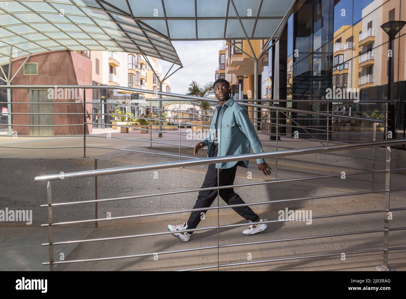 Side view of young black male millennial in trendy clothes and sneakers looking away while walking on ramp near entrance of modern building in city Stock Photo
