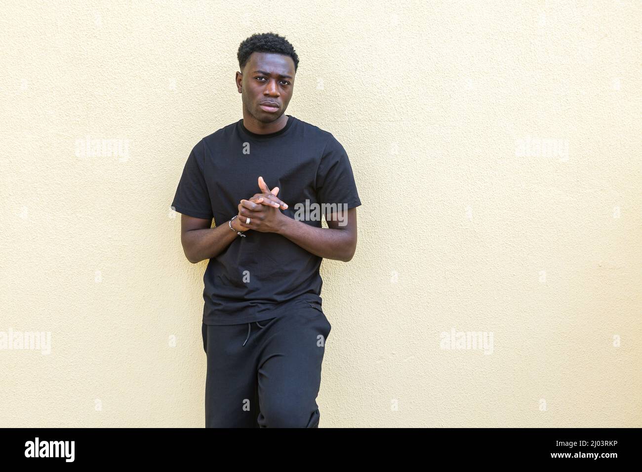 Self assured young African American male with dark hair in black casual wear standing near colored concrete wall and looking at camera Stock Photo