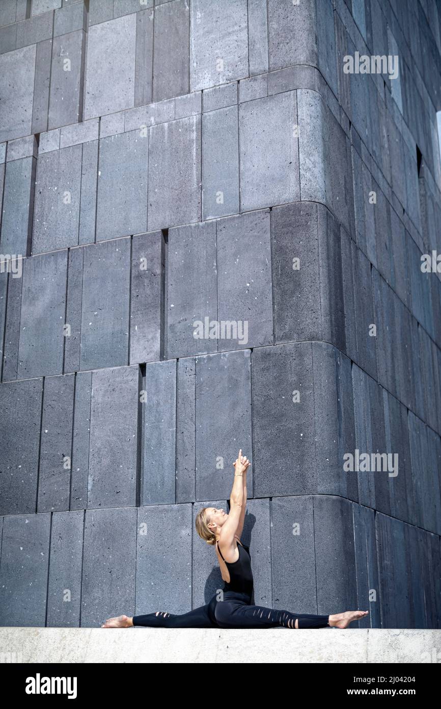 Charming and elegant woman practicing yoga in front of a concrete wall while doing a split Stock Photo