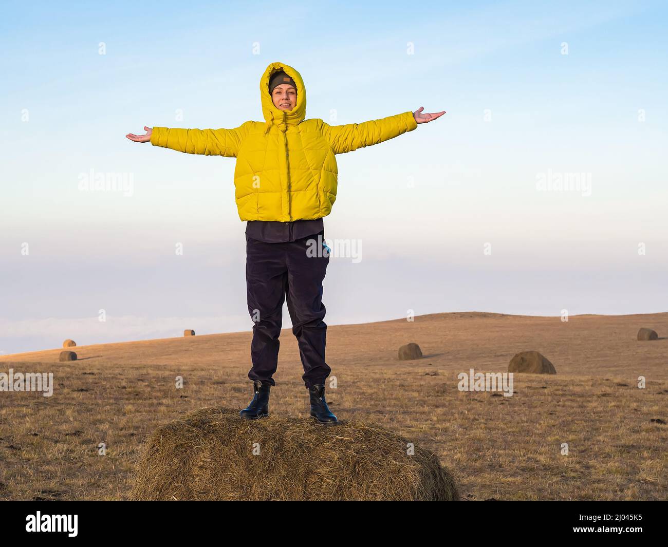 A girl in yellow jacket stands with her hands apart on a haystack in a