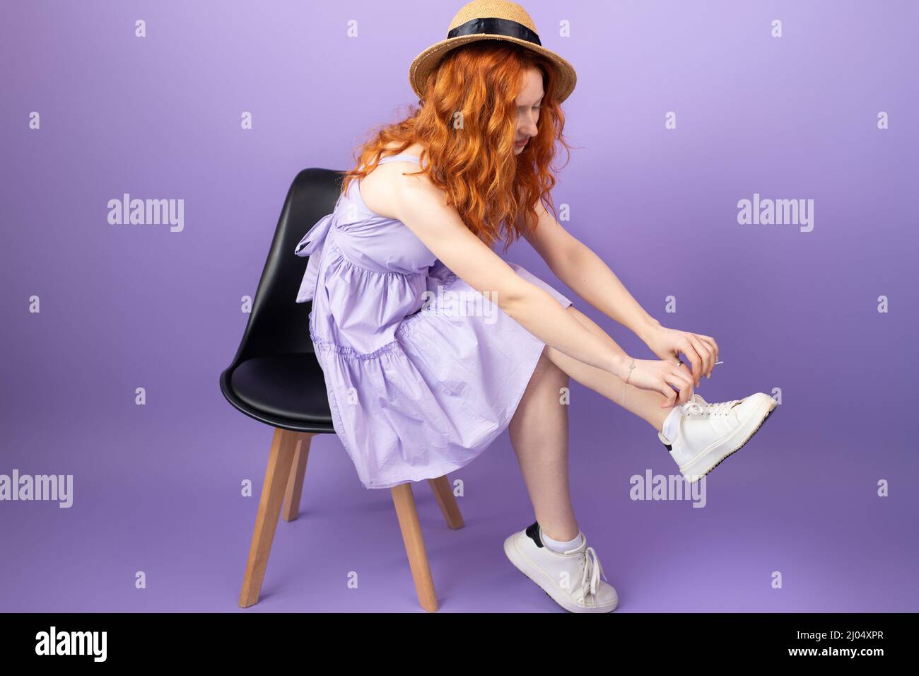 Full length body young redhead woman in straw hat and sundress sitting on the chair and tying shoelaces on white sneekers. Stock Photo