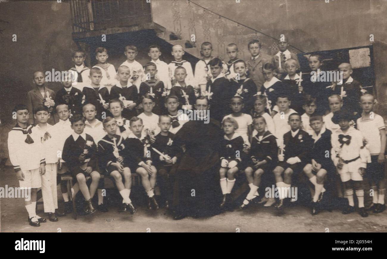 original antique 20th century photo about a boys  class in a hungarian primary school at the 1910s.with them teacher ( Head teacher) who was a roman catholic priest in usual uniform ( sitting in the first line and in the middle of the picture).  The photo was a big occasion at that time that's why all little boy are wearing them the nicest clothes and holding a candle in them hand. The traditional clothes was that time the sailor blouse in the Austro-Hungary empire ( nowadays tha sailor blouse for girls)  Period: 1910s Source: original photograph Stock Photo