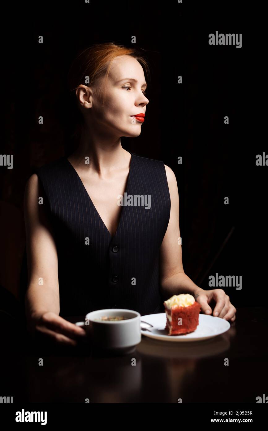 Woman at the table with tea and cake in the dark. Attractive forty-year-old woman in a black vest. Tea drinking. Loneliness. Stock Photo