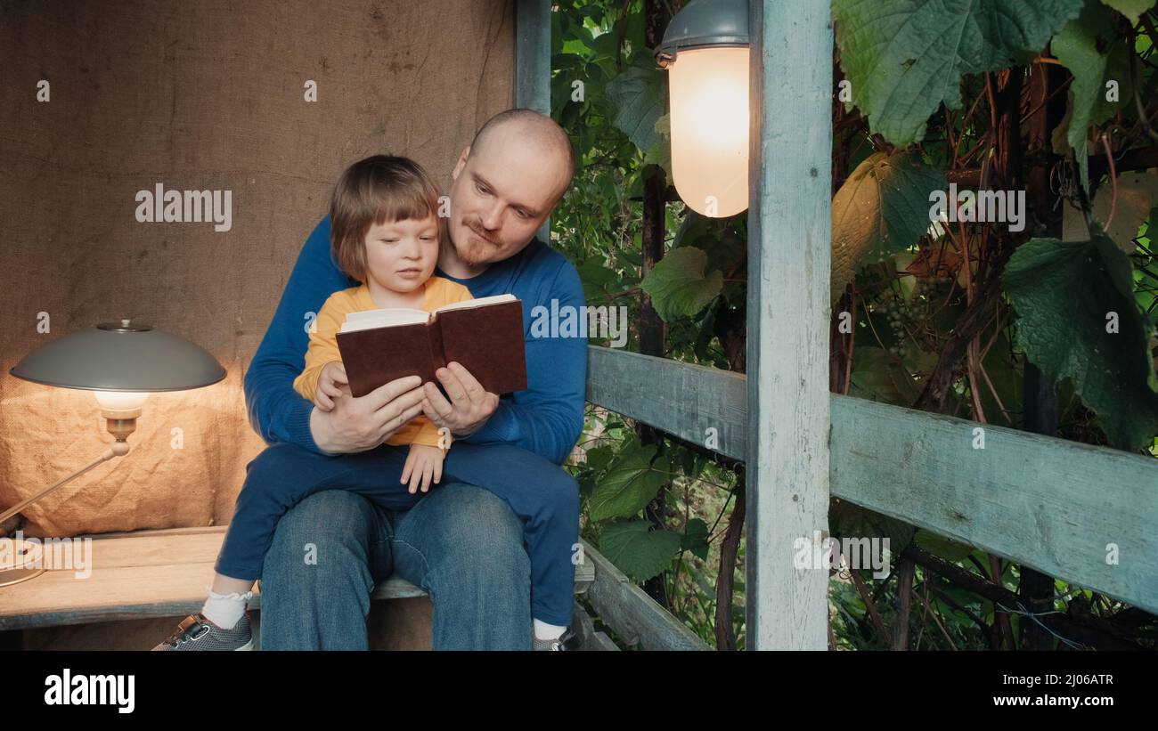 Man reads a paper book for small child, sitting on the porch of a village house Stock Photo