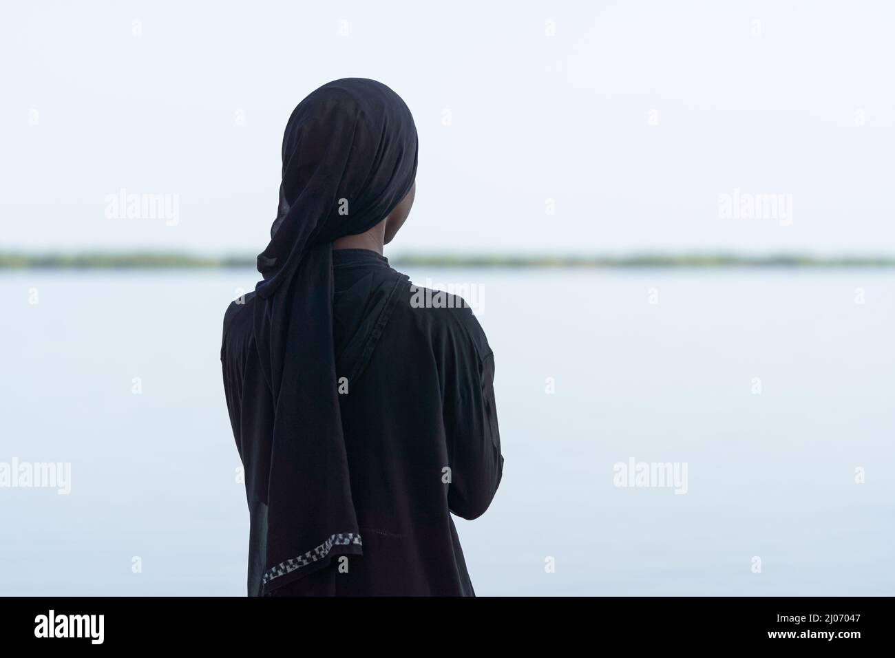 Rear view of an African girl dressed all in black looking over the sea and dreaming about her future; adolescent psychology and future orientation Stock Photo