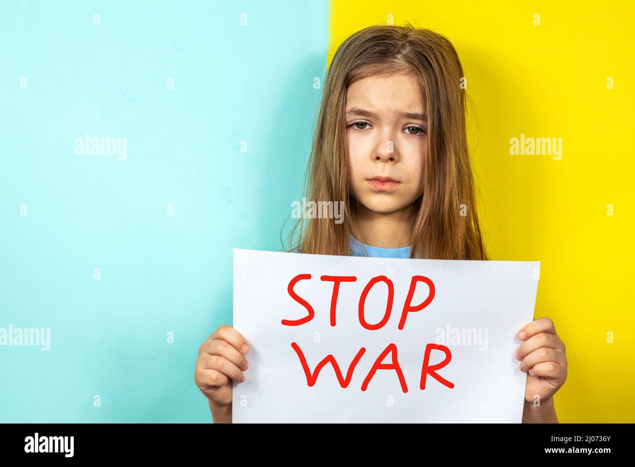 A girl is crying, holding a STOP WAR poster in her hands against the background of the Ukrainian flag. Stop the war in Ukraine. The concept of peace Stock Photo