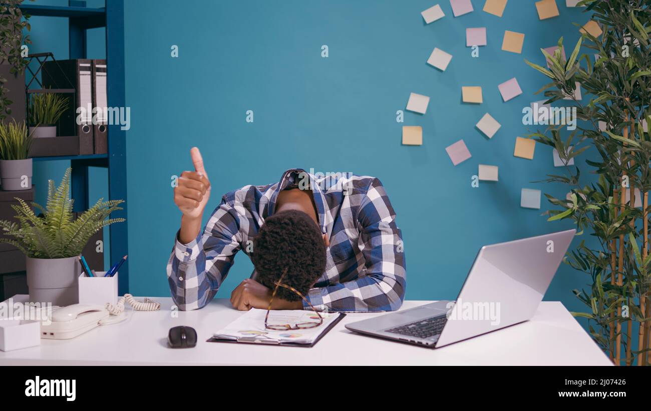 Tired employee resting head on desk and giving thumbs up in front of